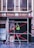 A person wearing a reflective safety vest stands on a ladder supported by a scaffold. The setting is an urban environment with a tiled floor. Behind, there is the exterior of a building with large windows reflecting intricate architectural details. Signs are present on the walls, and the ladder and scaffold assembly is secured with red crossbars.