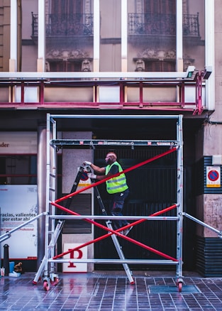 A person wearing a reflective safety vest stands on a ladder supported by a scaffold. The setting is an urban environment with a tiled floor. Behind, there is the exterior of a building with large windows reflecting intricate architectural details. Signs are present on the walls, and the ladder and scaffold assembly is secured with red crossbars.