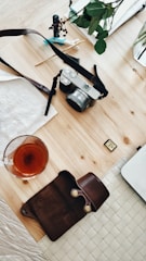 A flat lay of a wooden table featuring a camera with a strap, a dark leather pouch, a glass of tea, a small memory card, and a part of a laptop. There are some small toys or figurines and green leaves, adding a touch of playfulness and nature.