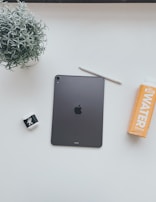 A minimalist arrangement on a white surface featuring a black iPad, a white stylus, a potted plant with green leaves, a pair of white AirPods with a black and white design on the case, and an orange water bottle labeled 'JUST WATER'.