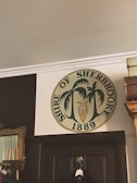 A circular emblem is mounted on an interior wall above a doorway. The emblem reads 'Shire of Sherbrooke 1889' and features palm tree graphics and a central design resembling an anchor or symbol. Nearby, there's a decorative mirror with an ornate frame, and a clothing rack is partially visible.