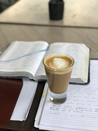 A glass of coffee with latte art rests on an open notebook filled with handwritten notes in Spanish. The scene appears to be set on a table with a blurred background, suggesting a calm and studious environment.