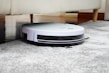 A robotic vacuum cleaner is navigating under a sofa on a gray carpet. The sleek design features a white top and black sides, with visible brushes peeking out underneath. The setting appears to be a living room with wooden flooring and a light-colored sofa.