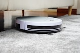 A robotic vacuum cleaner is navigating under a sofa on a gray carpet. The sleek design features a white top and black sides, with visible brushes peeking out underneath. The setting appears to be a living room with wooden flooring and a light-colored sofa.