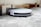 A robotic vacuum cleaner is navigating under a sofa on a gray carpet. The sleek design features a white top and black sides, with visible brushes peeking out underneath. The setting appears to be a living room with wooden flooring and a light-colored sofa.