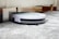 A robotic vacuum cleaner is navigating under a sofa on a gray carpet. The sleek design features a white top and black sides, with visible brushes peeking out underneath. The setting appears to be a living room with wooden flooring and a light-colored sofa.