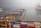 A shipping port filled with colorful stacked cargo containers lined up in rows. Several large cranes are positioned along the docks, ready for loading and unloading. Trucks and equipment are visible, indicating a bustling hub of activity despite the overcast weather. The water appears calm with ships docked in the background.