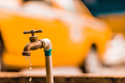 A close-up view of a brass water faucet with droplets of water falling from it. The background features an out-of-focus orange vehicle, creating a vibrant contrast.