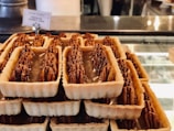 A display of rectangular tart pastries filled with pecans, neatly arranged on a glass counter. These sweet treats have a golden-brown crust and are topped with glossy pecan halves. The background shows a blurred café interior with a label indicating the type of pastry.