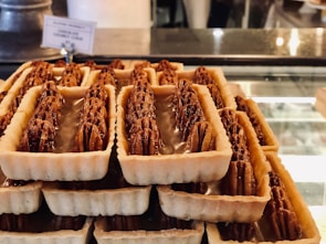 A display of rectangular tart pastries filled with pecans, neatly arranged on a glass counter. These sweet treats have a golden-brown crust and are topped with glossy pecan halves. The background shows a blurred café interior with a label indicating the type of pastry.
