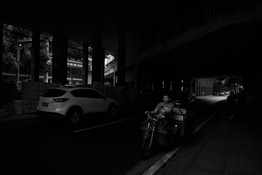 A dimly lit urban street scene under an overpass where a man is riding a motorized tricycle laden with goods. A white SUV is parked on the left, and a few people walk along the sidewalk. The image captures the contrast between the darker foreground and the lighted background of the street.