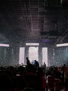 A crowded concert venue with people gathered in front of a brightly lit stage. Confetti is falling from above, adding to the festive atmosphere. Silhouetted figures can be seen amongst the audience, some holding up phones to capture the performance. The stage is framed by large rectangular light panels.