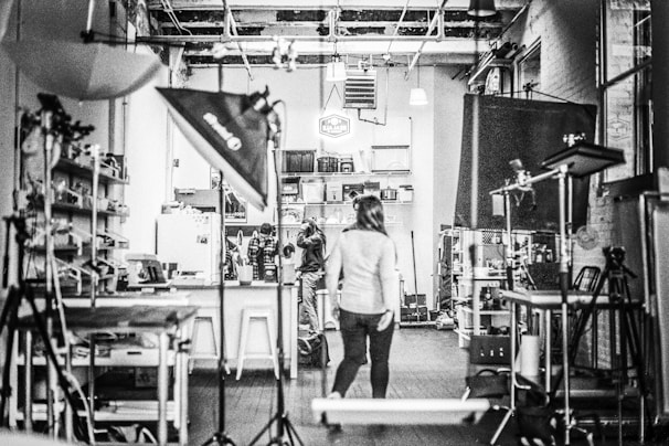 A black and white image of a studio with equipment like lights and stands scattered around. Shelving units and a variety of equipment are visible, suggesting a creative or photographic workspace. Three people are located centrally, one walking towards the back where a cluttered area is filled with boxes and materials.