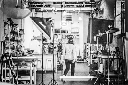A black and white image of a studio with equipment like lights and stands scattered around. Shelving units and a variety of equipment are visible, suggesting a creative or photographic workspace. Three people are located centrally, one walking towards the back where a cluttered area is filled with boxes and materials.