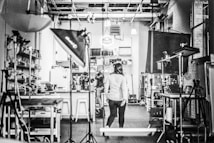A black and white image of a studio with equipment like lights and stands scattered around. Shelving units and a variety of equipment are visible, suggesting a creative or photographic workspace. Three people are located centrally, one walking towards the back where a cluttered area is filled with boxes and materials.