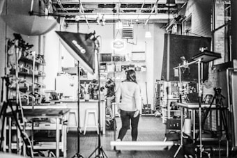A black and white image of a studio with equipment like lights and stands scattered around. Shelving units and a variety of equipment are visible, suggesting a creative or photographic workspace. Three people are located centrally, one walking towards the back where a cluttered area is filled with boxes and materials.
