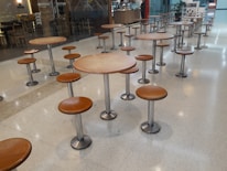A seating area with multiple round tables and attached stools, all featuring metal bases and brown wooden tops. The tables are arranged in neat rows on a polished light-colored floor. In the background, there is a counter or small cafe with some signage and a couple of people.