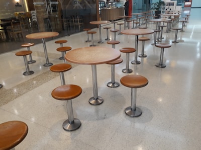 A seating area with multiple round tables and attached stools, all featuring metal bases and brown wooden tops. The tables are arranged in neat rows on a polished light-colored floor. In the background, there is a counter or small cafe with some signage and a couple of people.