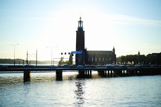 A silhouette of a large building with a tall central tower, likely a city hall, is positioned beside a bridge. The structure is reflected in the water below, creating a serene waterfront scene. Streetlights and light traffic are visible on the bridge, with trees in the background under a clear sky at sunset or sunrise.