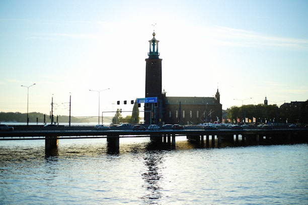 A silhouette of a large building with a tall central tower, likely a city hall, is positioned beside a bridge. The structure is reflected in the water below, creating a serene waterfront scene. Streetlights and light traffic are visible on the bridge, with trees in the background under a clear sky at sunset or sunrise.