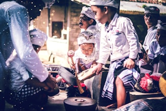 A group of children wearing traditional attire are gathered together, intently playing musical instruments. The scene is vibrant with a focus on cultural expression, as each child is engaged in creating music. The light and shadows add a dramatic effect to their focused expressions and movements.