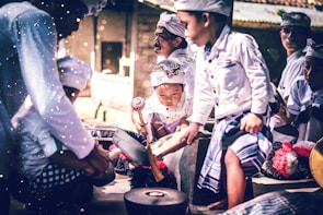 A group of children wearing traditional attire are gathered together, intently playing musical instruments. The scene is vibrant with a focus on cultural expression, as each child is engaged in creating music. The light and shadows add a dramatic effect to their focused expressions and movements.