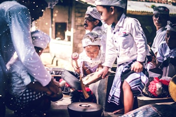 A group of children wearing traditional attire are gathered together, intently playing musical instruments. The scene is vibrant with a focus on cultural expression, as each child is engaged in creating music. The light and shadows add a dramatic effect to their focused expressions and movements.