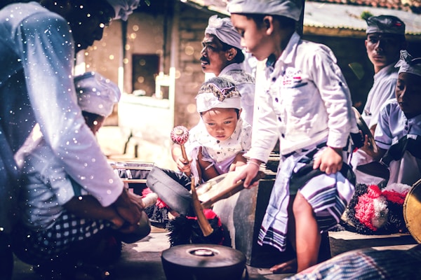 A group of children wearing traditional attire are gathered together, intently playing musical instruments. The scene is vibrant with a focus on cultural expression, as each child is engaged in creating music. The light and shadows add a dramatic effect to their focused expressions and movements.