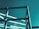 A partially constructed steel framework structure against a solid teal-colored sky. The metal beams intersect at various angles, forming geometric patterns and suggesting an industrial or architectural setting.