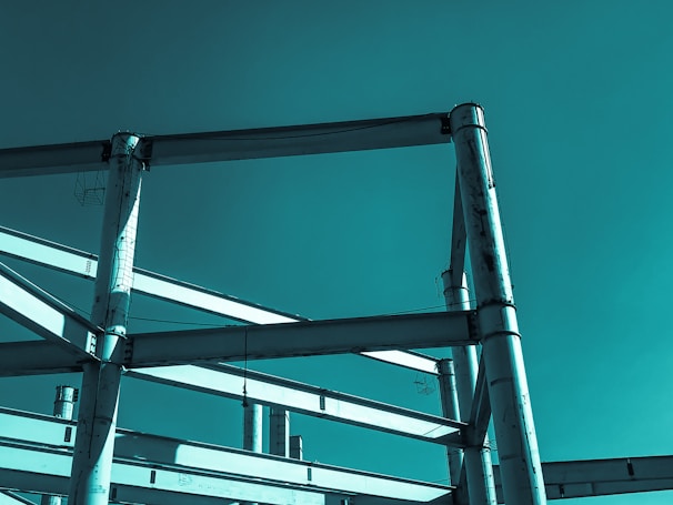 A partially constructed steel framework structure against a solid teal-colored sky. The metal beams intersect at various angles, forming geometric patterns and suggesting an industrial or architectural setting.
