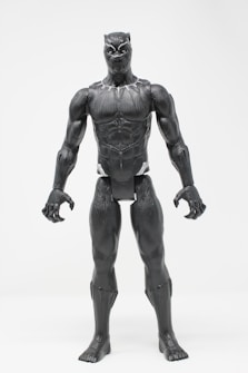 A detailed action figure of a superhero character standing against a plain white background. The figure is highly detailed with a muscular build and textured black suit, featuring a distinctive superhero mask with pointed ears and sleek design elements.