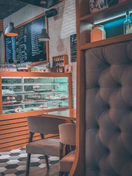 A cozy café interior featuring a display case filled with various pastries and confectioneries. The setting includes a tufted seating area, wooden accents, and modern pendant lighting. A blackboard menu with handwritten items is visible, and shelves hold decorative jars and other items.