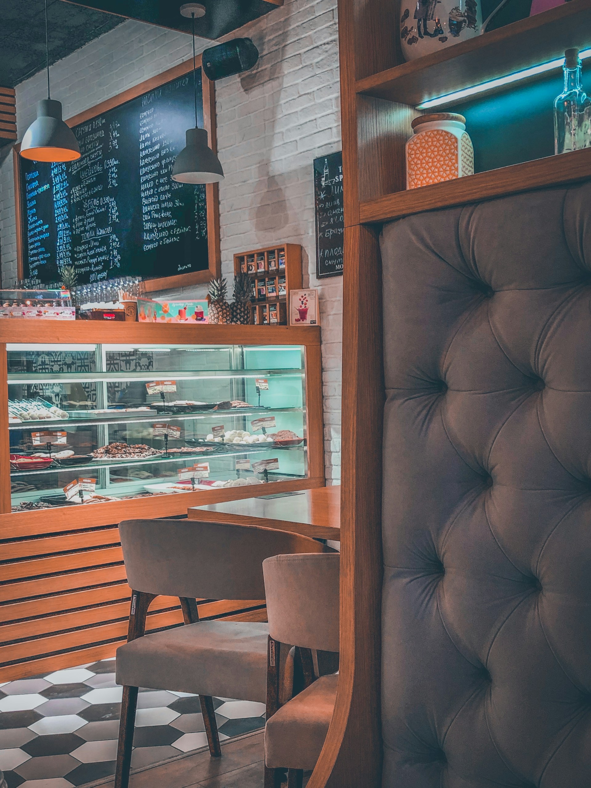 A cozy café interior featuring a display case filled with various pastries and confectioneries. The setting includes a tufted seating area, wooden accents, and modern pendant lighting. A blackboard menu with handwritten items is visible, and shelves hold decorative jars and other items.