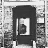 A historic architectural structure with a rustic brick exterior. The entrance leads to a view of a hallway through a uniquely shaped window. A sign with text is posted on the wall next to the entrance.