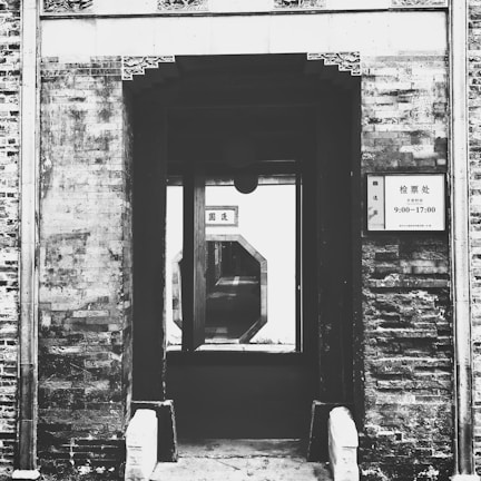 A historic architectural structure with a rustic brick exterior. The entrance leads to a view of a hallway through a uniquely shaped window. A sign with text is posted on the wall next to the entrance.
