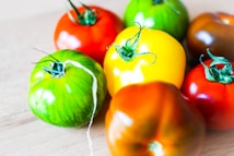 A close-up view of several heirloom tomatoes of varying colors, including red, green, yellow, and orange, arranged on a light wooden surface. The tomatoes have a shiny texture and their stems are visible, adding a fresh and vibrant feel.