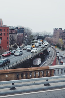 Heavy traffic is observed on a multi-lane urban highway surrounded by brick and concrete buildings. Cars and trucks are positioned bumper-to-bumper, suggesting congestion. Trees without leaves are visible, hinting at a winter season.