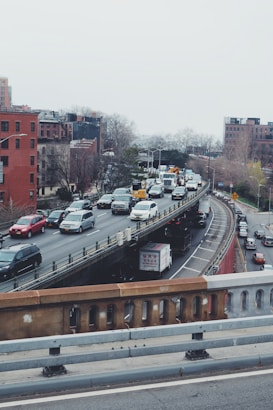 Heavy traffic is observed on a multi-lane urban highway surrounded by brick and concrete buildings. Cars and trucks are positioned bumper-to-bumper, suggesting congestion. Trees without leaves are visible, hinting at a winter season.
