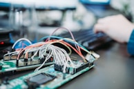 A close-up of a circuit board with numerous wires attached, showcasing an intricate setup of electronic components. In the background, a person is typing on a keyboard, indicating a work environment involving electronics or programming.