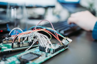 A close-up of a circuit board with numerous wires attached, showcasing an intricate setup of electronic components. In the background, a person is typing on a keyboard, indicating a work environment involving electronics or programming.