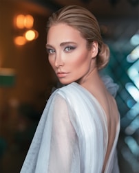 A woman with styled blonde hair gazes confidently over her shoulder while wearing a sheer, elegant top. The setting is softly lit with warm lights creating a blurred bokeh effect in the background, while cool tones enhance the overall mood.