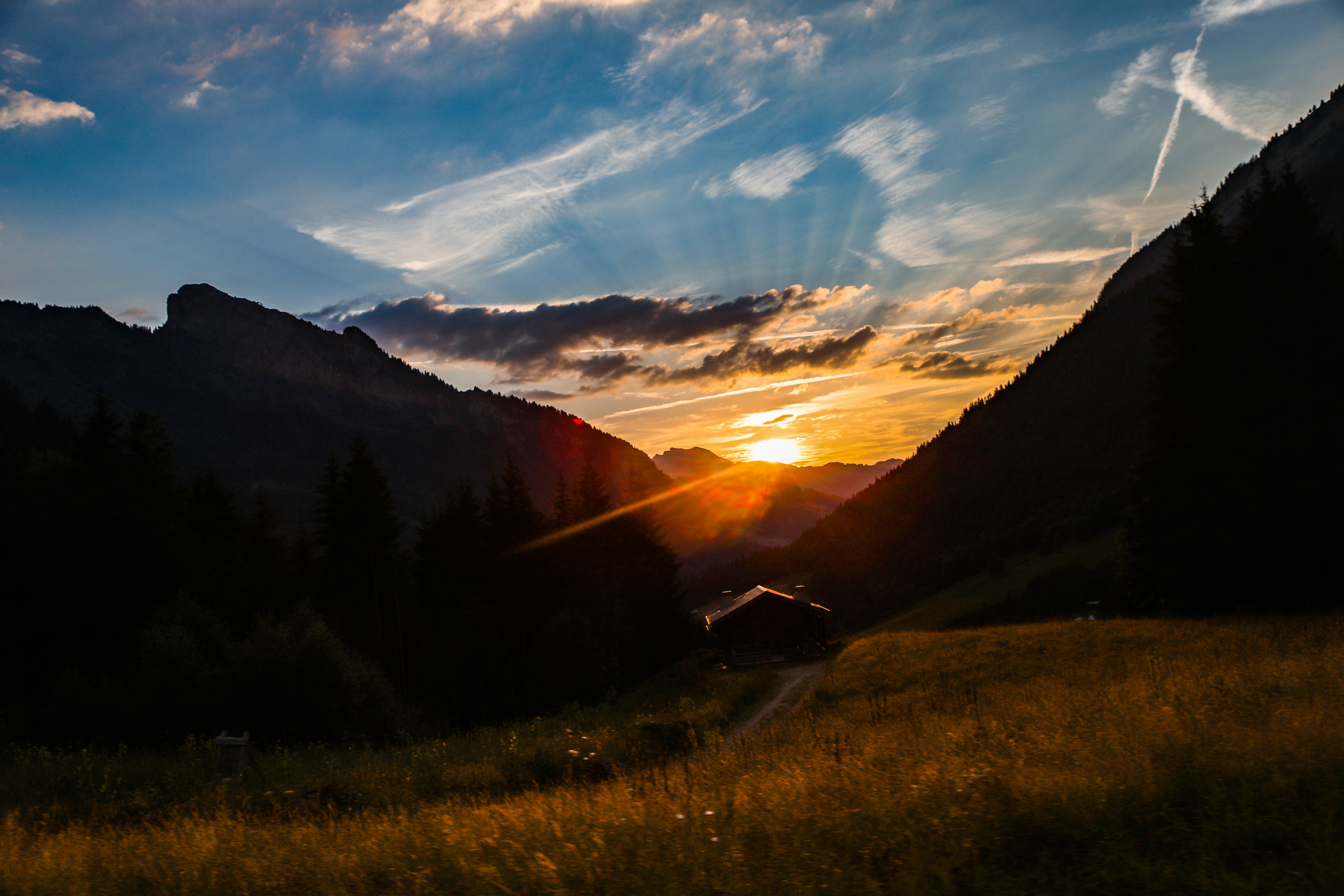 At the Lac des Mines d'Or, on the French Alps, the sun sets between two mountains, behind the idyllic scene of a simple wooden house, with a long path in the foreground. The field to the front creates an air of romance and happiness.