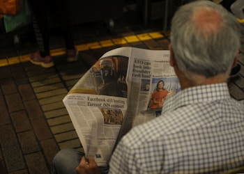 An older man with gray hair, wearing a checkered shirt, is sitting and reading a newspaper. The newspaper features several articles with different headlines and images. The setting appears to be indoors, with brick flooring and another person standing nearby, partially visible.