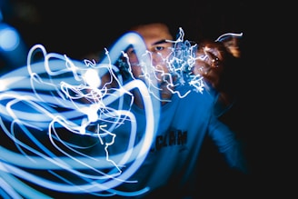 A person is partly visible, surrounded by a chaotic swirl of bright, blue and white light trails against a dark background. The light gives a dynamic and energetic feel, almost as if the person is manipulating electricity.