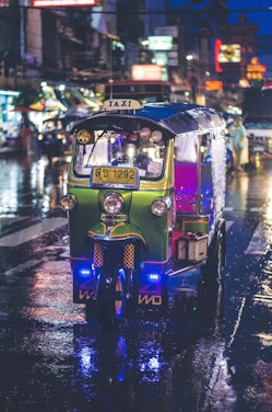 A brightly colored tuk-tuk is driving on a rain-soaked street at night. Neon lights and reflections create a vibrant ambiance, with blurred city lights in the background.