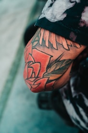 A close-up image of an arm with a colorful tattoo featuring a design that includes large red flowers and bold black outlines. The tattoo appears to extend across the elbow area, and the person's clothing is partially visible with a patterned, tie-dye-like shirt.