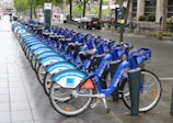 A row of blue rental bicycles is neatly lined up on a wet sidewalk in an urban setting. The bikes have advertisements on their frames, and there is a bike docking station visible. Street signs, trees, parked cars, and pedestrians can be seen in the background, suggesting a city environment.