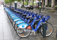 A row of blue rental bicycles is neatly lined up on a wet sidewalk in an urban setting. The bikes have advertisements on their frames, and there is a bike docking station visible. Street signs, trees, parked cars, and pedestrians can be seen in the background, suggesting a city environment.