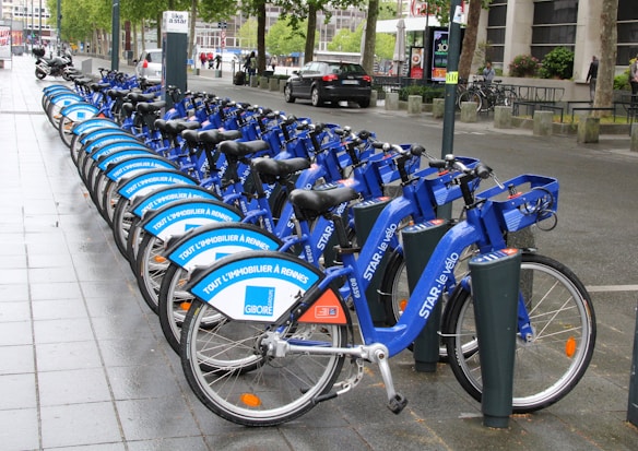 A row of blue rental bicycles is neatly lined up on a wet sidewalk in an urban setting. The bikes have advertisements on their frames, and there is a bike docking station visible. Street signs, trees, parked cars, and pedestrians can be seen in the background, suggesting a city environment.
