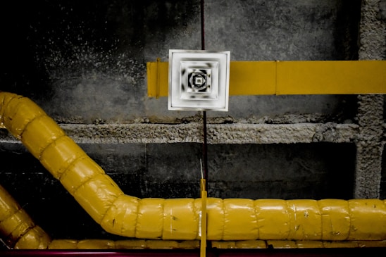 A series of interconnected yellow ventilation pipes against a rough concrete ceiling. A central square vent with a blurred, black and white pattern is positioned between the pipes and the ceiling.